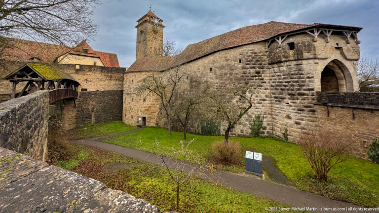Large stone gate block and defensive wall with a tall tower behind and a covered timber passage crossing the left side.