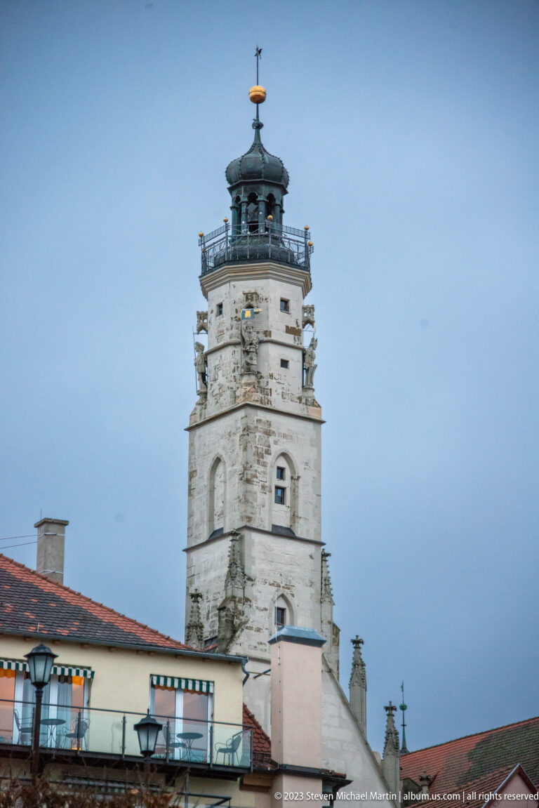 White weathered tower with arched openings, projecting upper gallery, dark domed cupola, and weather vane above tiled roofs.