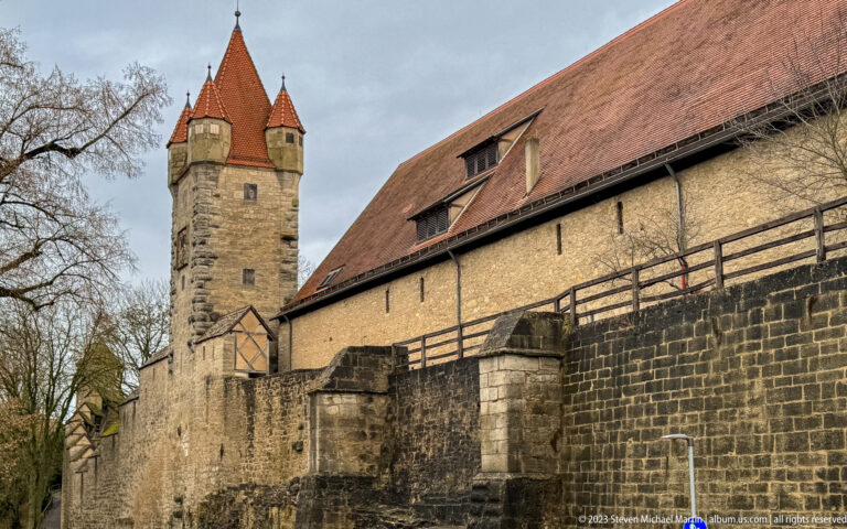 Long roofed stone fortification wall with timber gallery and a tall roofed defensive tower rising at the left.