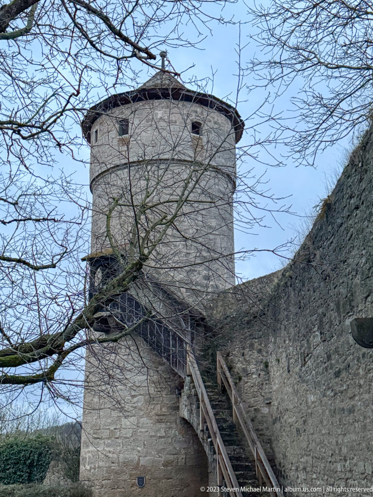 Round stone defensive tower joined to the city wall by a steep wooden stair, with bare branches crossing the view.