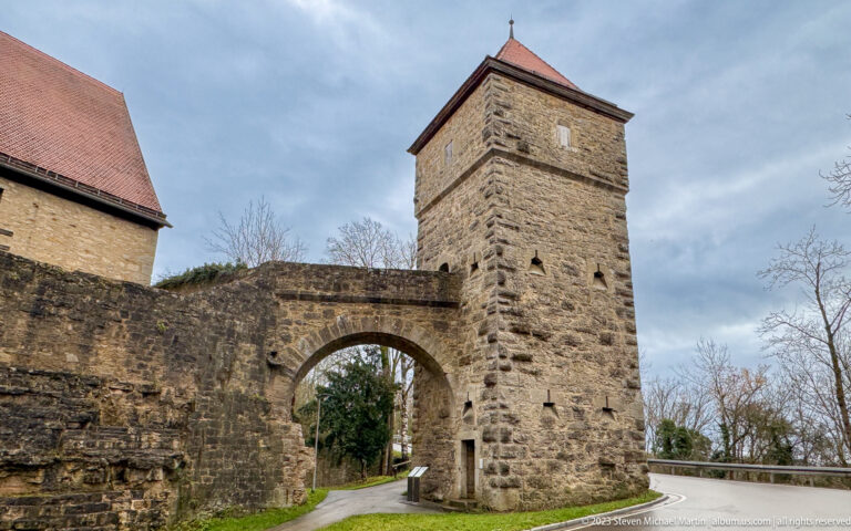 Square stone gate tower with broad arched passage, small slit openings, red-tiled roof, and adjoining wall beside a curving road.