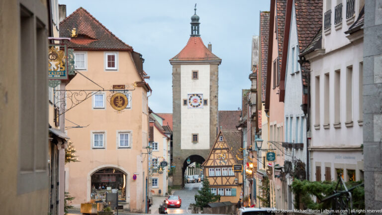 Historic street lined with plastered and half-timbered houses leading to a pale clock-faced gate tower with an arched passage.