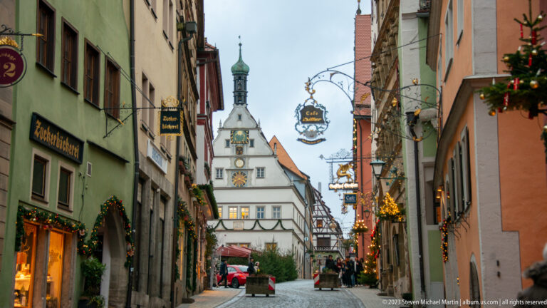 Decorated historic street leading toward the white gabled Ratstrinkstube with clock displays on Rothenburg’s Marktplatz.