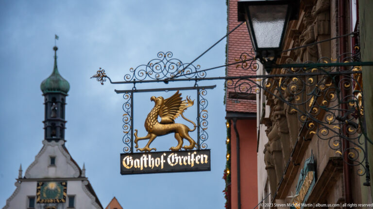 Ornate wrought-iron Gasthof Greifen sign with a gilded winged griffin, with the Ratstrinkstube tower blurred behind.