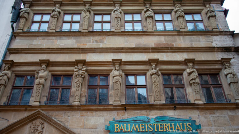 Upper façade of the Baumeisterhaus with rows of windows, carved standing figures, stone cornices, and a turquoise name sign.