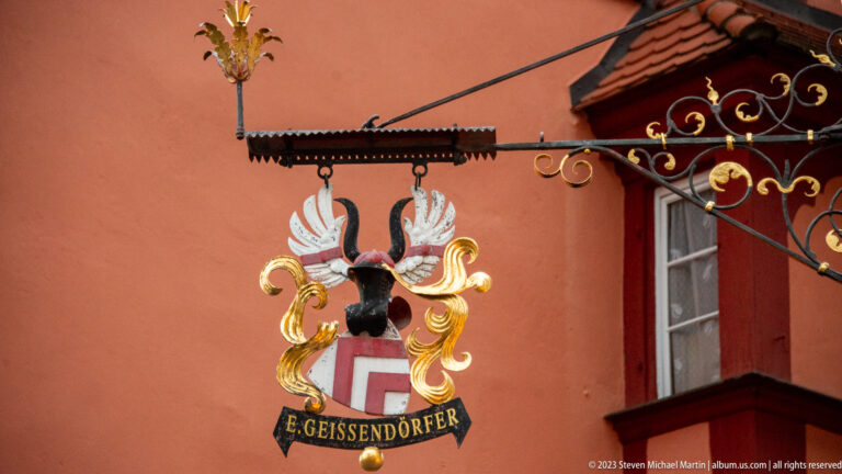 Painted heraldic sign reading “E. Geissendörfer,” with shield, helmet crest, and white-and-gold mantling on a red wall.