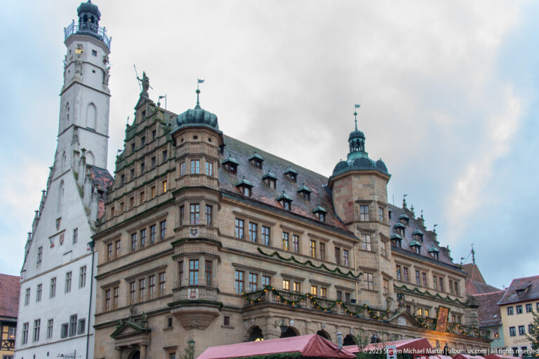 Rothenburg Town Hall with arcades, dormered roof, projecting corner tower, stepped gable, and Christmas-market stalls in front.