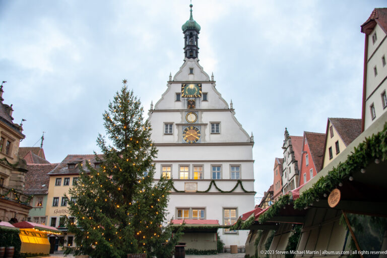 Large lit Christmas tree on Marktplatz in front of the white gabled Ratstrinkstube and surrounding market stalls.
