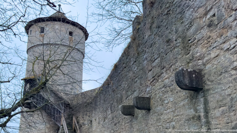 Close view of a rough stone city wall with projecting blocks, a round defensive tower, and timber stair at left.