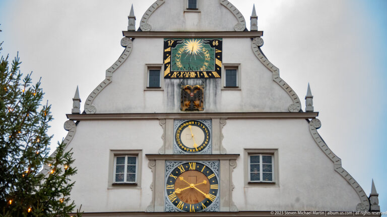 White Ratstrinkstube gable with large lower clock, smaller circular dial, colorful sun-and-zodiac display, and pointed finials.