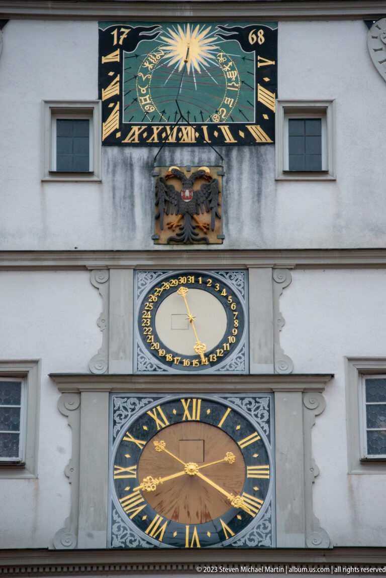 Close detail of the Ratstrinkstube’s colorful celestial panel, heraldic eagle, small dial, and large Roman-numeral clock.