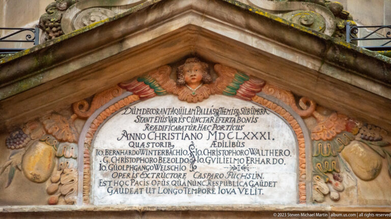 Close view of a Latin inscription tablet dated in Roman numerals, surrounded by carved fruit, scrolls, masks, and a cherub head.