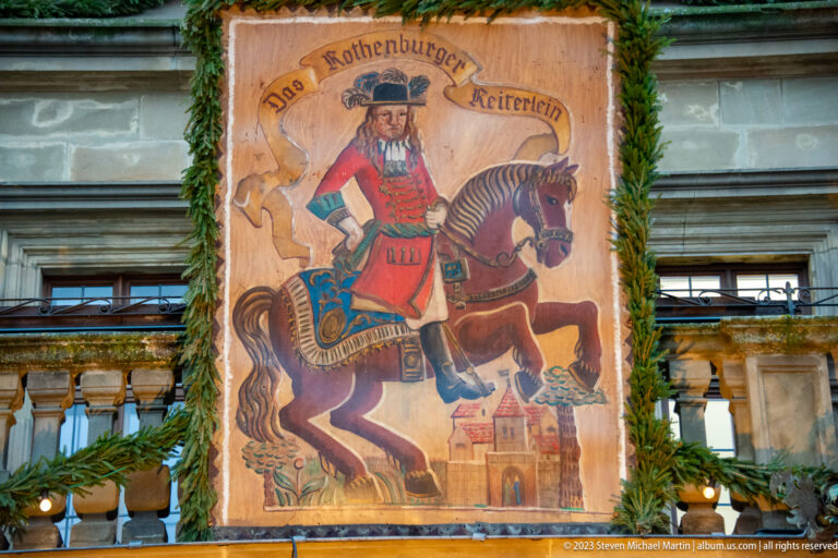 Painted banner reading “Das Rothenburger Reiterlein,” with a costumed rider on horseback above a small town scene and Christmas greenery.