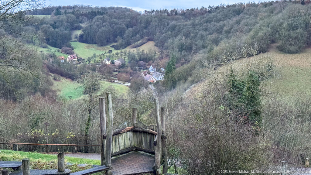 Rustic wooden footbridge overlooking green fields, scattered houses, orchards, and wooded slopes in the Tauber valley.