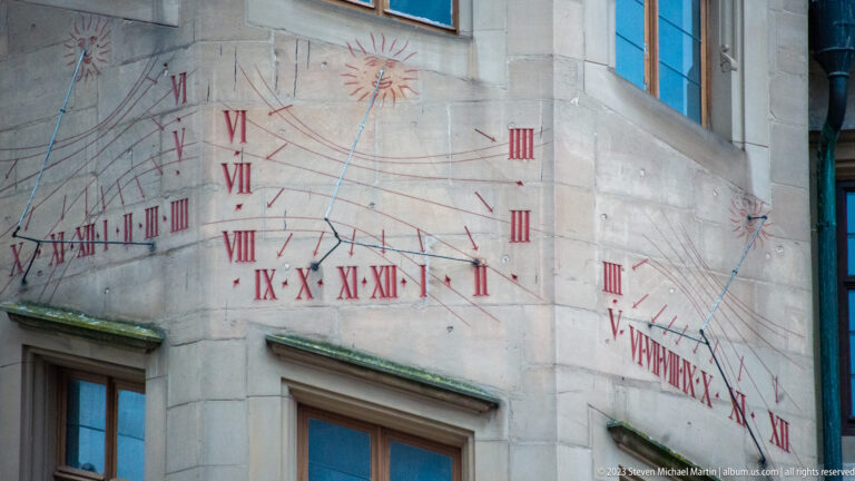 Painted red sundial wrapping around a stone building corner with Roman numerals, hour lines, sun motifs, and multiple gnomons.