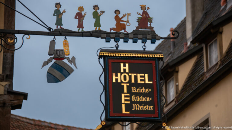 Illuminated hanging sign reading “Hotel Reichsküchenmeister,” suspended from ornate wrought iron with small silhouetted figures.
