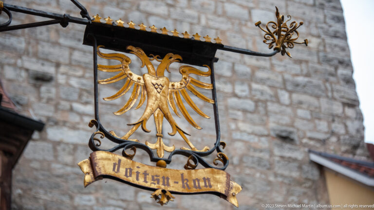 Black hanging sign with a gilded double-headed eagle and ribbon inscription, supported by ornate wrought-iron scrollwork.