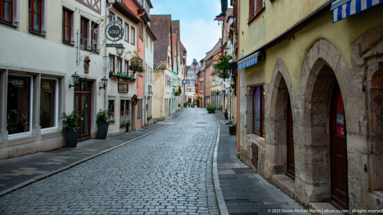 Quiet cobblestone street lined with shops, historic façades, stone arcades, hanging signs, flower boxes, and seasonal greenery.