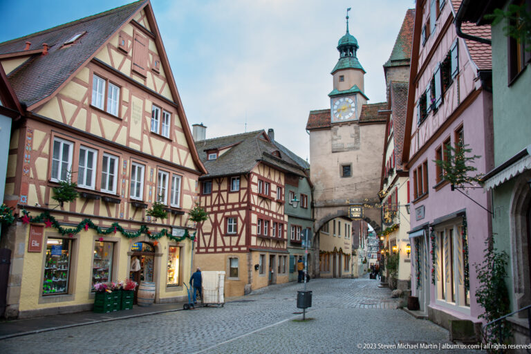 Broad cobblestone street with colorful half-timbered houses leading to Markusturm, its clock face and green turret centered above the arch.