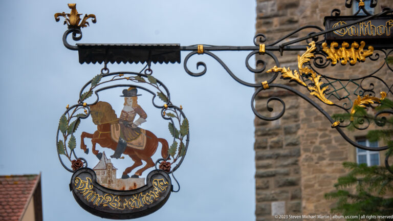 Ornate circular wrought-iron sign with a mounted rider and curved name panel, set against a rough stone tower wall.