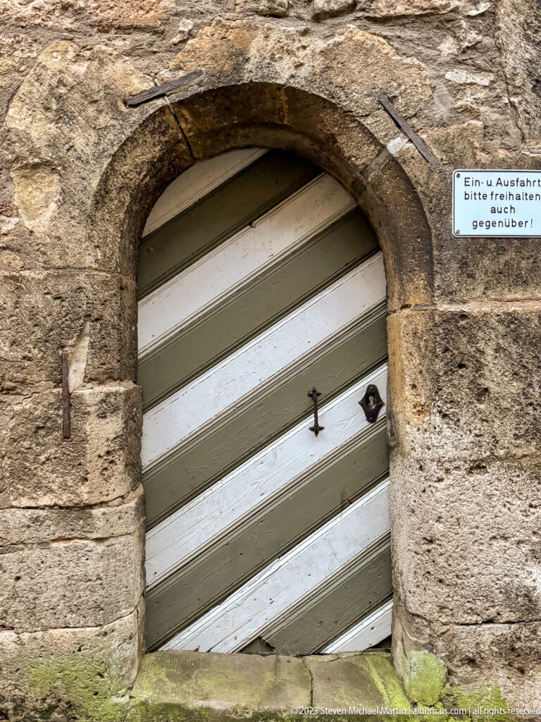 Low stone arch containing a diagonally boarded striped timber door with iron fittings and a small German warning sign.