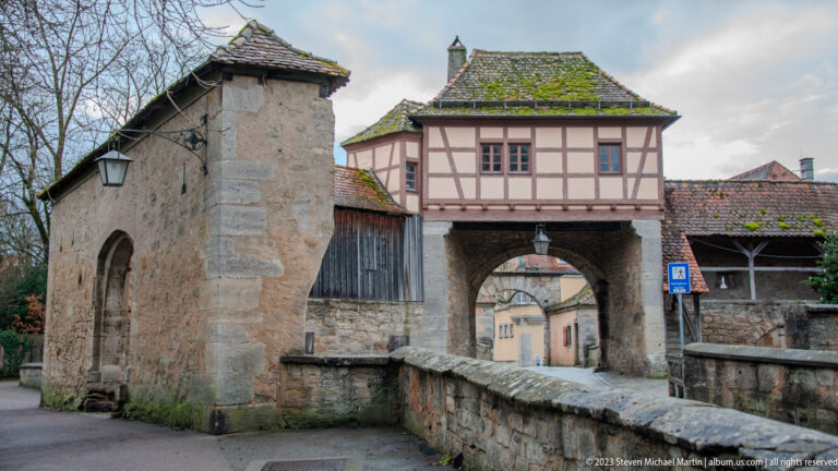 Half-timbered upper story over a stone arched gateway, with rough walls, mossy tiled roofs, and adjoining timber structures.