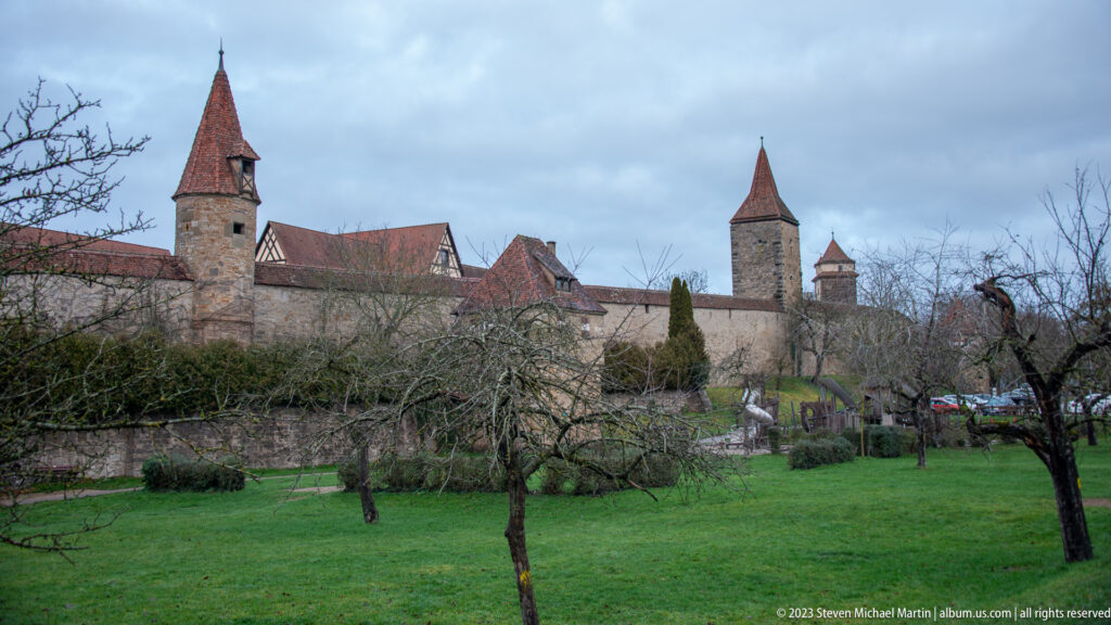 Long stone city wall with several roofed defensive towers rising beyond a grassy orchard with bare fruit trees.