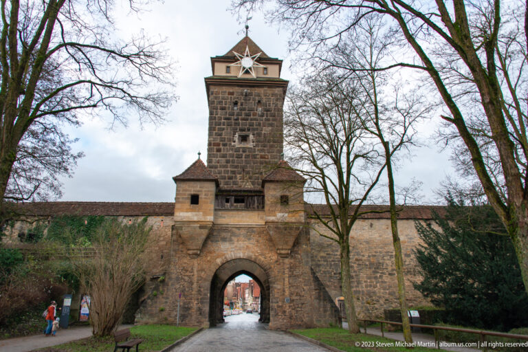 Square stone gate tower with broad arch, small flanking structures, shallow roof, cupola, and a large star decoration.