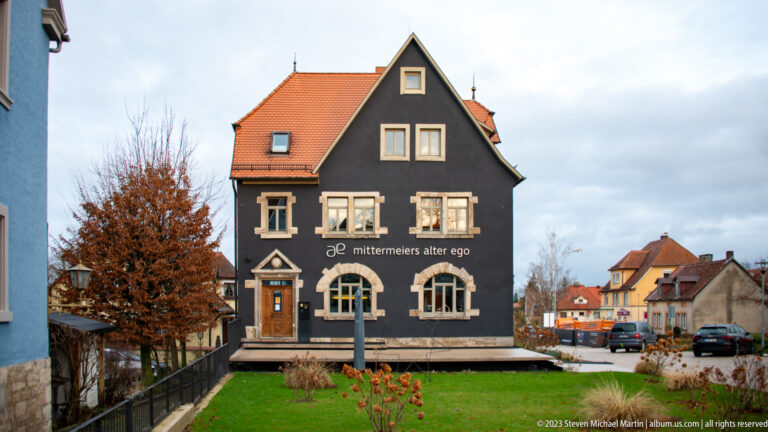 Dark gabled building marked “mittermeiers alter ego,” with orange tile roof, pale window surrounds, arched windows, and front lawn.