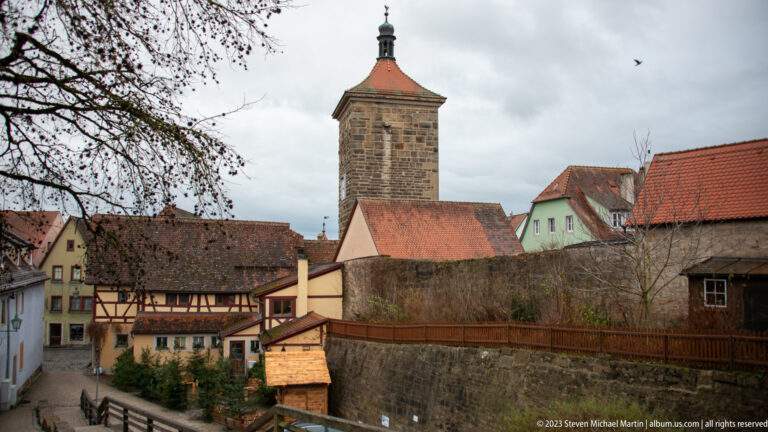 Tall square stone tower above red-roofed and half-timbered houses, with a high masonry wall across the foreground.