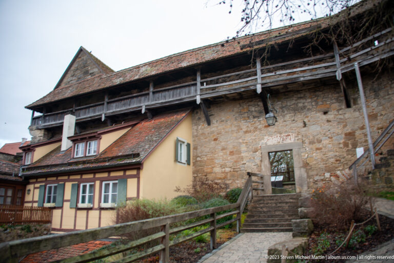 Stone stair beside a half-timbered house leading to a timber-supported covered wall walk along the massive city wall.
