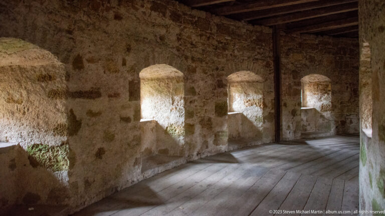 Interior of a covered wall walk with three deep stone embrasures, narrow openings, timber ceiling beams, and wooden floor.