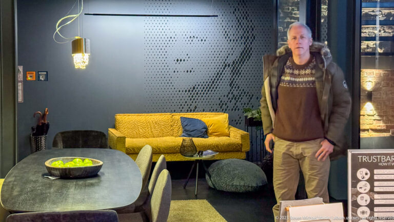 Modern guest-room lounge with exposed brick, yellow sofa, dining table, and a man standing beside the seating area.
