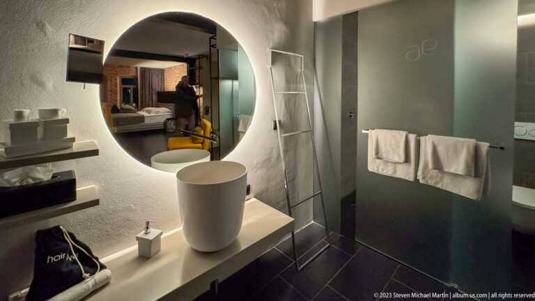 Backlit round mirror and white vessel sink beside a glass shower enclosure with two hanging towels.