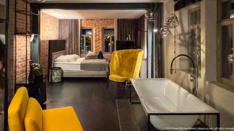 Long view through a guest suite with bed, exposed brick, yellow chairs, and a freestanding white bathtub.