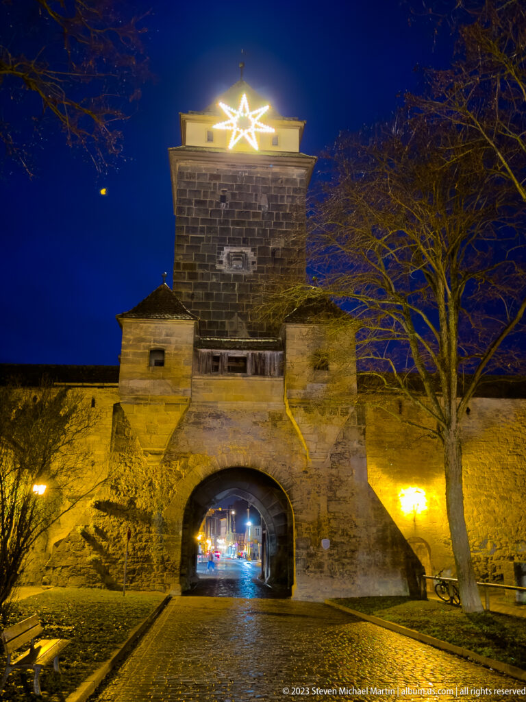 Floodlit medieval city gate with square tower, arched passage, stone walls, and a glowing Christmas star above.