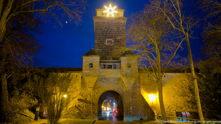 Wide night view of a square medieval gate tower, adjoining walls, bare trees, and a bright star above.