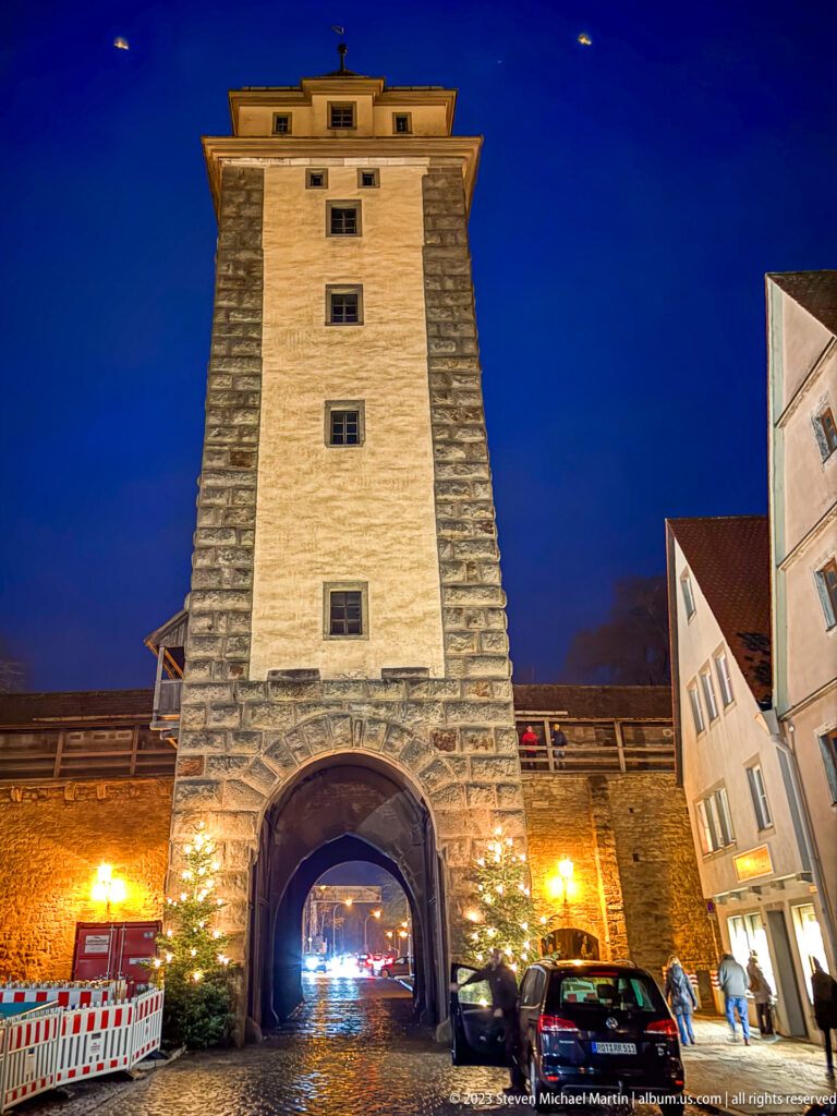 Rectangular medieval gate tower with arched passage, rough stone edges, parked cars, and illuminated wall at night.