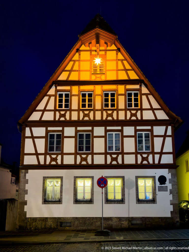 Gabled half-timbered house with white infill, dark timber framing, lit windows, and a glowing star near the roof peak.