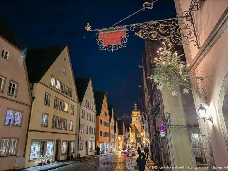 Narrow night street lined with gabled houses, shop windows, pedestrians, and an ornate wrought-iron hanging sign.