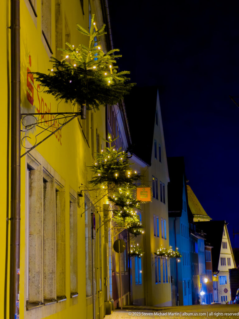 Historic street façade decorated with small lit evergreen arrangements, with gabled houses receding into the night.