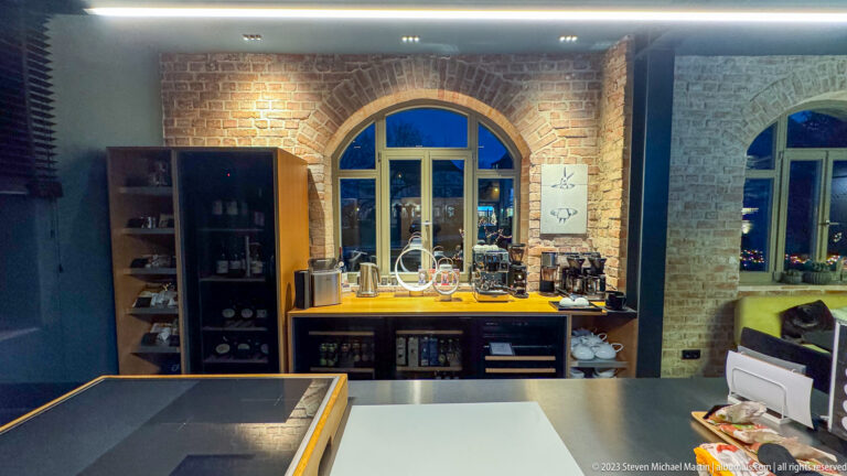 Brick-arched hotel kitchen area with counters, shelving, glassware, and coffee equipment beneath a rounded window.