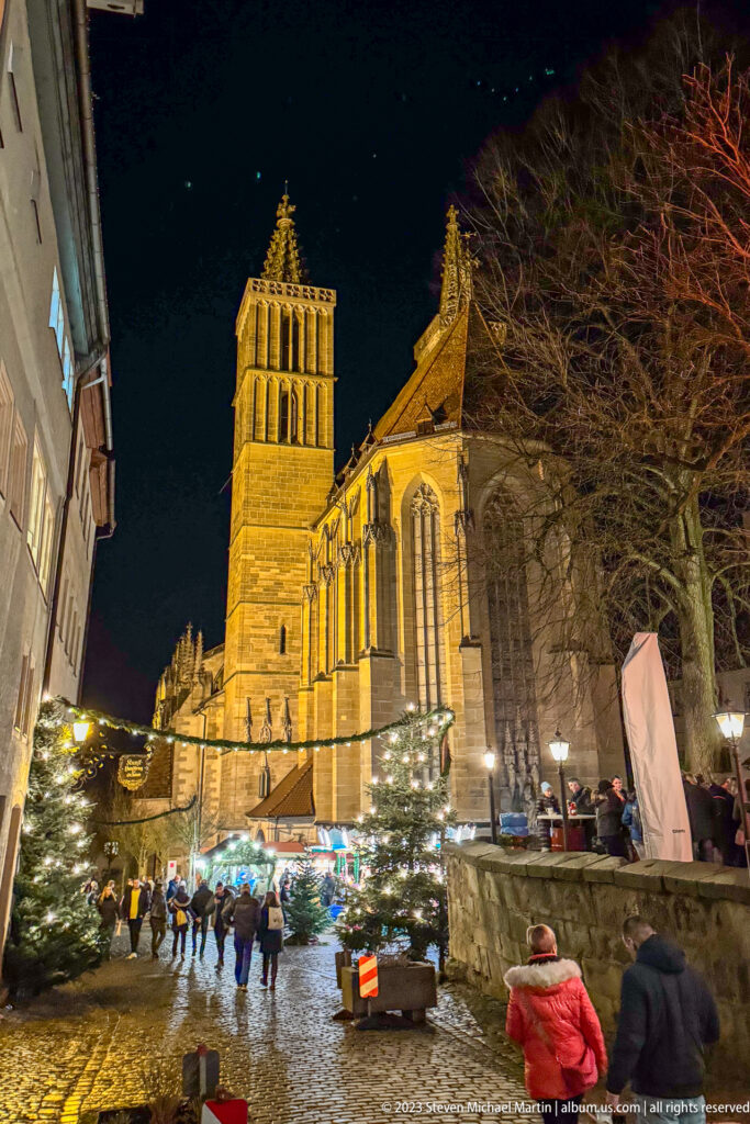 Illuminated Gothic St. James's Church above Christmas market stalls, small trees, and pedestrians on a wet street.