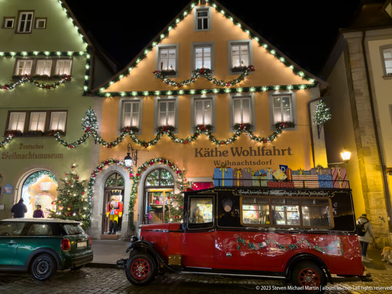Illuminated Käthe Wohlfahrt and German Christmas Museum façade with garlands, lights, a Christmas tree, and a red vintage vehicle.
