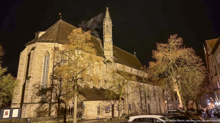 Floodlit St. Wolfgang's Church with Gothic windows, buttresses, steep roof, and slender turret behind bare winter trees.