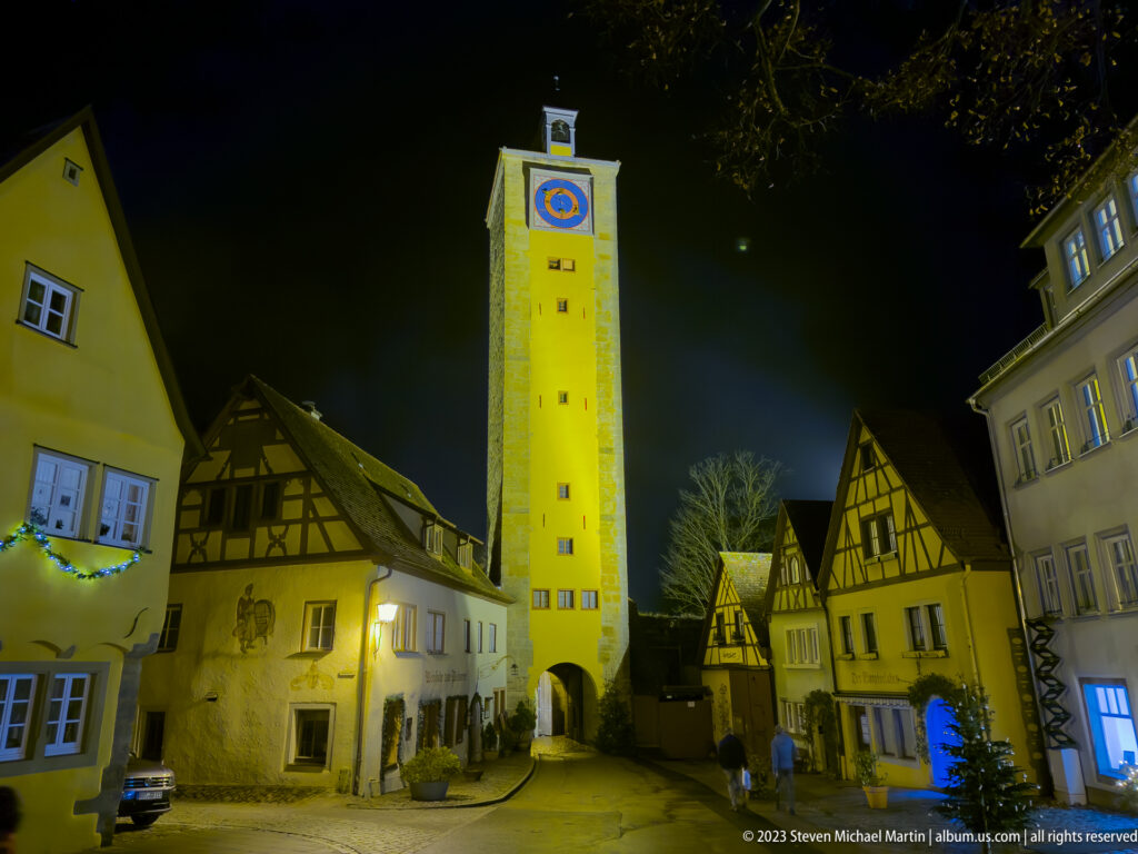 Illuminated Klingentorturm with clock face and arched gateway rising above half-timbered houses at night.