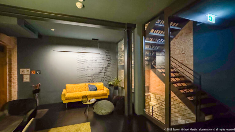 Split-level room with exposed brick, open metal staircase, yellow sofa, and dark modern lounge furniture.