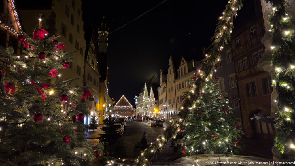 Christmas trees with red ornaments and white lights frame a gabled old-town street leading toward brighter buildings at night.