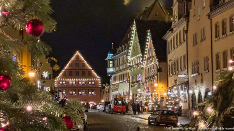 Christmas-lit historic street framed by decorated evergreens, with gabled buildings, pedestrians, and parked cars.