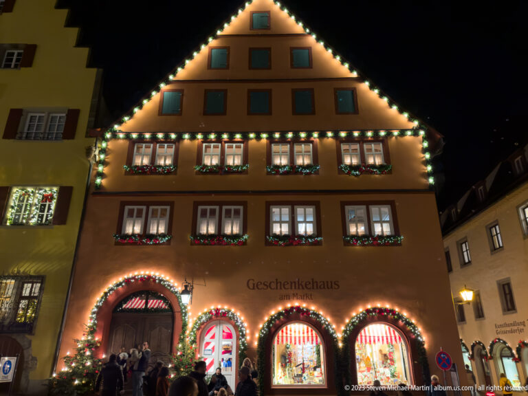 Illuminated gabled Geschenkehaus am Markt façade with garlands, arched shop windows, and visitors below.